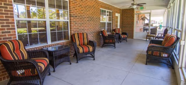 A covered outdoor patio area with several wicker chairs featuring red and orange striped cushions, and matching wicker tables. The patio has a concrete floor, brick walls with windows, and a ceiling fan. The area is enclosed on one side with large windows and has an exit door at the far end.