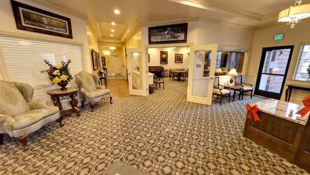 Interior view of a senior living facility lobby area with patterned carpet, two upholstered armchairs and a small round table with a floral arrangement on the left, a hallway leading to other rooms, and a seating area with chairs and tables visible through open double doors. There is a reception desk with holiday decorations on the right near an exit door with windows.
