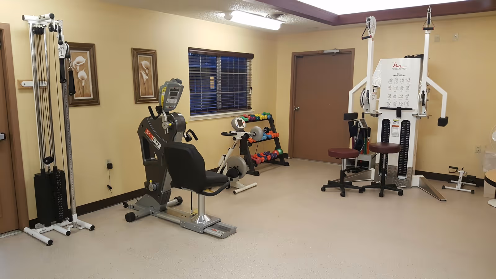 A small exercise room with various fitness equipment including a recumbent exercise bike, a multi-station weight machine, a stationary bike, and a rack of colorful dumbbells. The room has beige walls, two framed floral pictures, a window with blinds, and two closed brown doors.