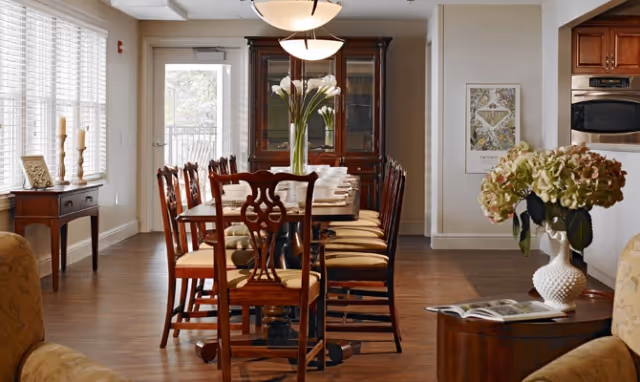 Long communal dining room with a wooden table and chairs, a china cabinet, pendant lights, hardwood floors, and floral centerpieces.