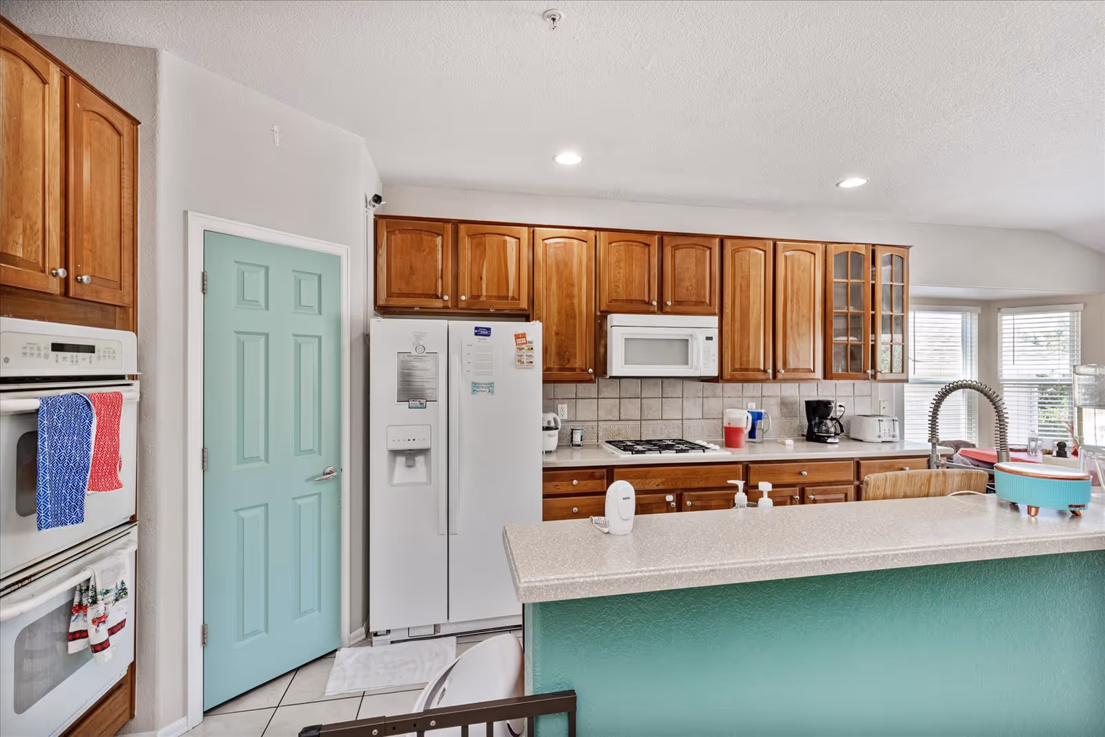 A bright kitchen with wooden cabinets, a white refrigerator, a built-in microwave, and a gas stove. There is a light green door and a matching green kitchen island with a beige countertop. Various kitchen appliances and items are on the counter, including a coffee maker, toaster, and hand soap dispensers. A window with blinds allows natural light into the space.