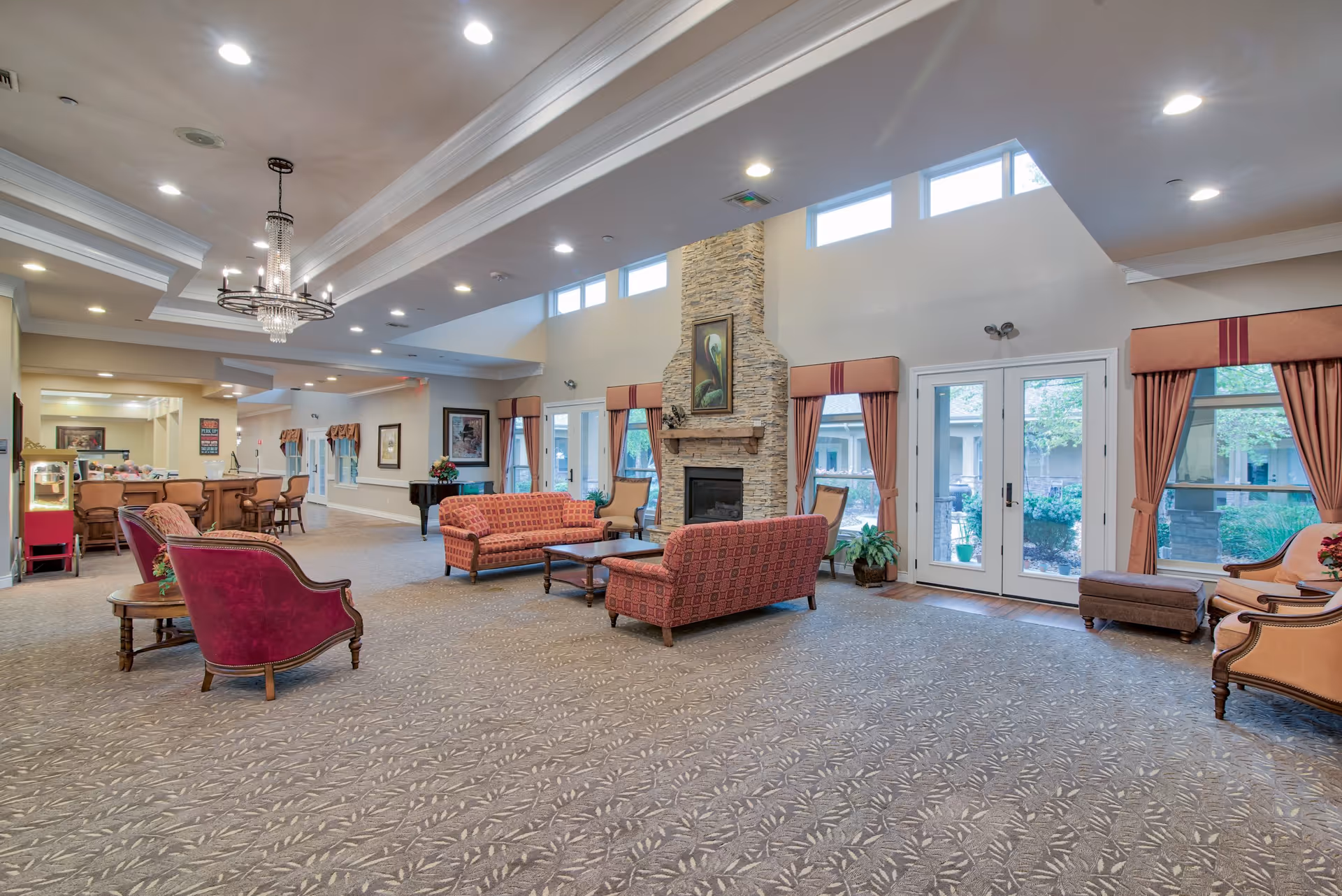Spacious and well-lit living room area in Pelican Bay Assisted Living Community featuring multiple upholstered sofas and chairs arranged around a stone fireplace with a painting above it. Large windows and glass doors with curtains allow natural light to fill the room, and a chandelier hangs from the ceiling. The room has carpeted flooring and a cozy, inviting atmosphere.