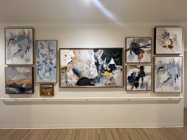 A wall decorated with a collection of nine abstract paintings in various sizes, featuring a mix of muted and dark colors. Below the paintings is a white handrail attached to the wall, and the floor is wooden.