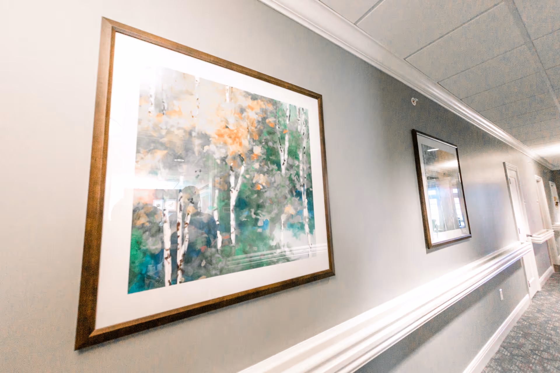 A hallway in a senior living facility with framed artwork on the walls. The walls are painted gray with white trim, and the floor is carpeted with a patterned design. The ceiling has recessed lighting and ceiling tiles.
