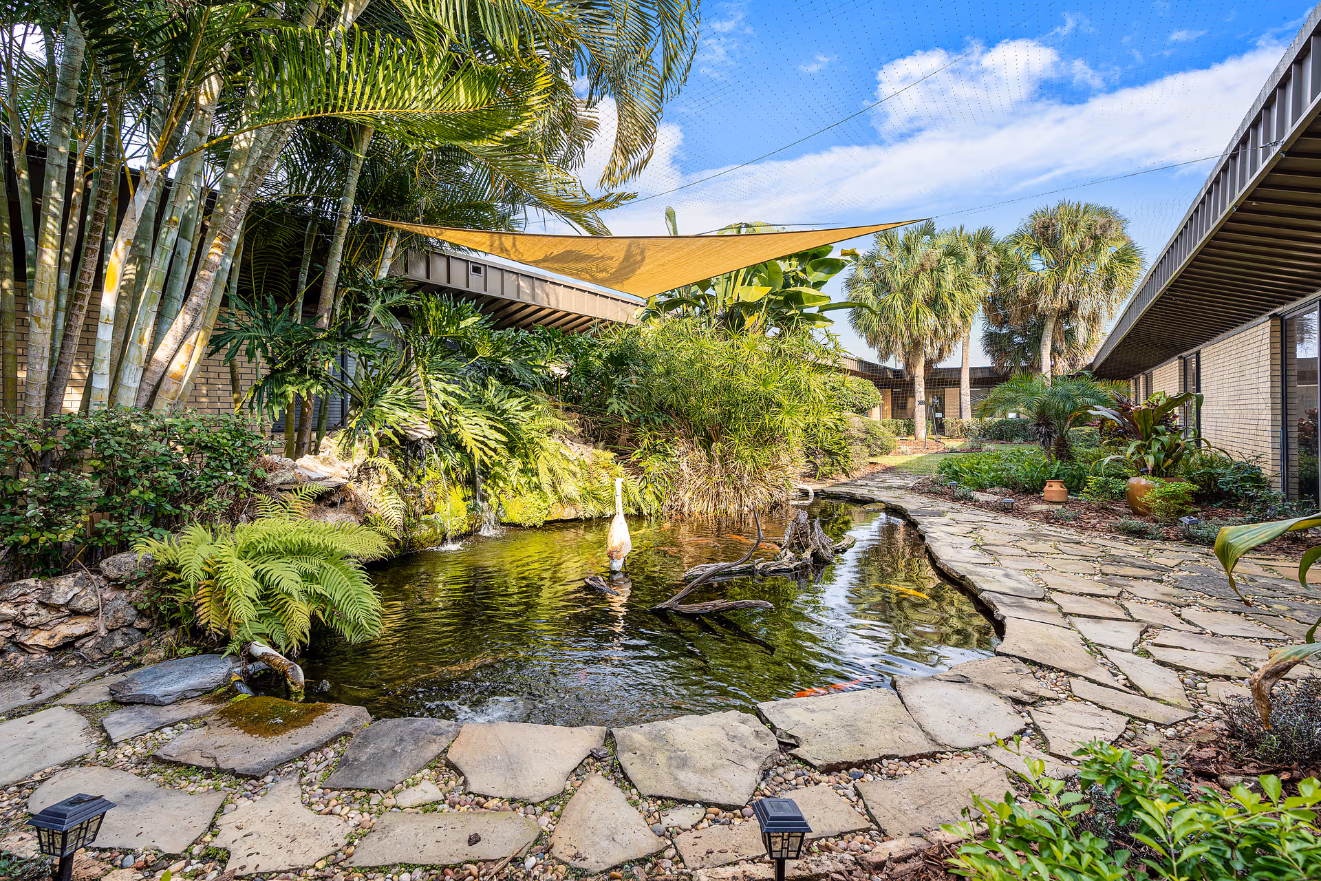 A tranquil outdoor garden area at Winter Garden Rehabilitation and Nursing Center featuring a small pond with a water fountain, surrounded by lush tropical plants and palm trees. Stone pathways border the pond, and a yellow triangular shade sail is stretched overhead. The building's exterior walls and windows are visible on both sides of the garden.