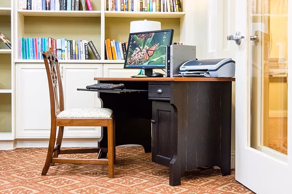 A small office area with a wooden desk and chair on a patterned carpet. The desk has a computer monitor displaying a butterfly on flowers, a keyboard, a mouse, and a printer. Behind the desk is a white built-in bookshelf filled with books. A glass door is partially visible on the right side.