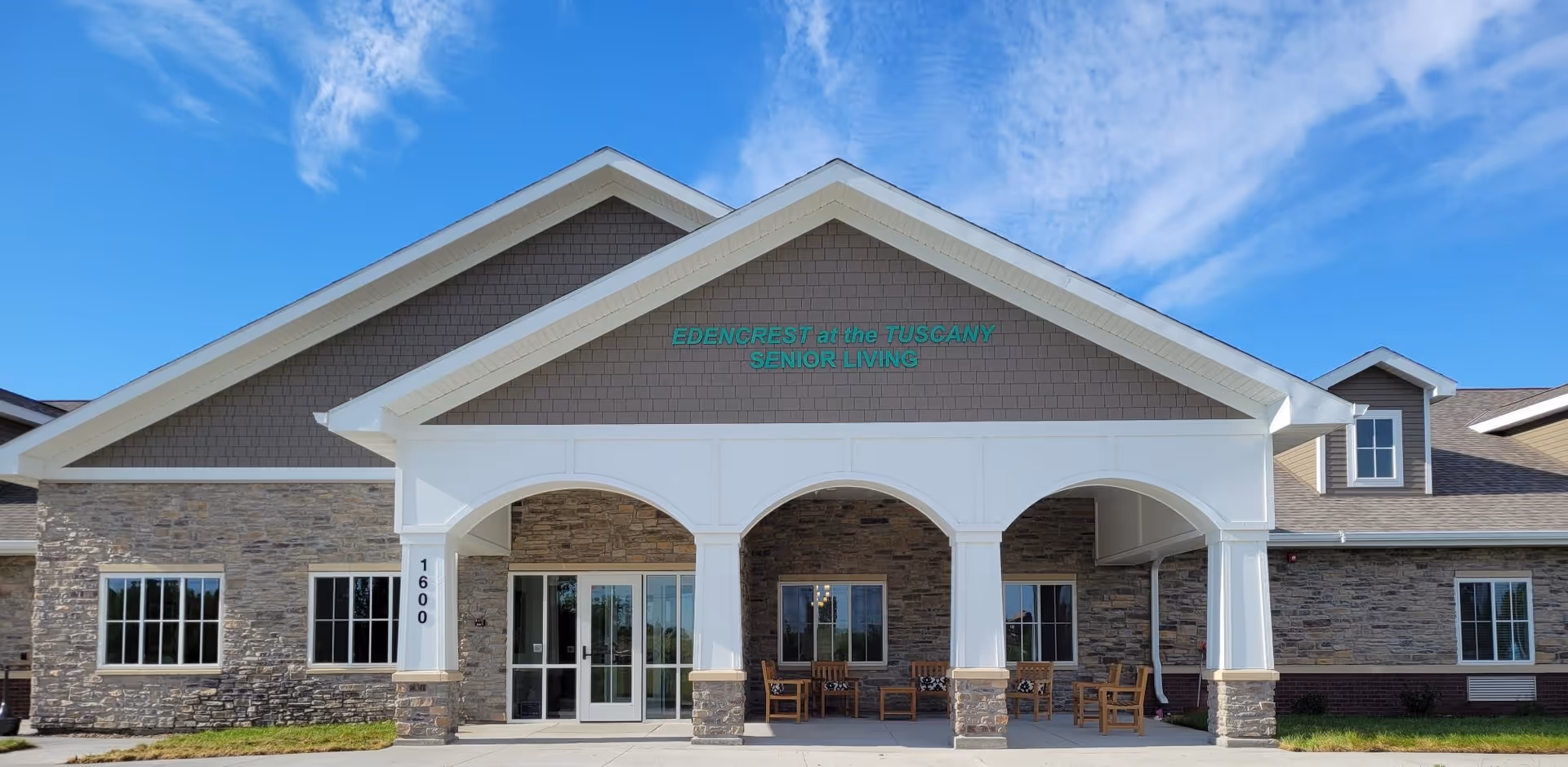 Front exterior view of Edencrest at The Tuscany senior living facility with stone and siding facade, large covered entrance supported by white columns, glass double doors, and outdoor seating under the porch. Blue sky with some clouds is visible above.