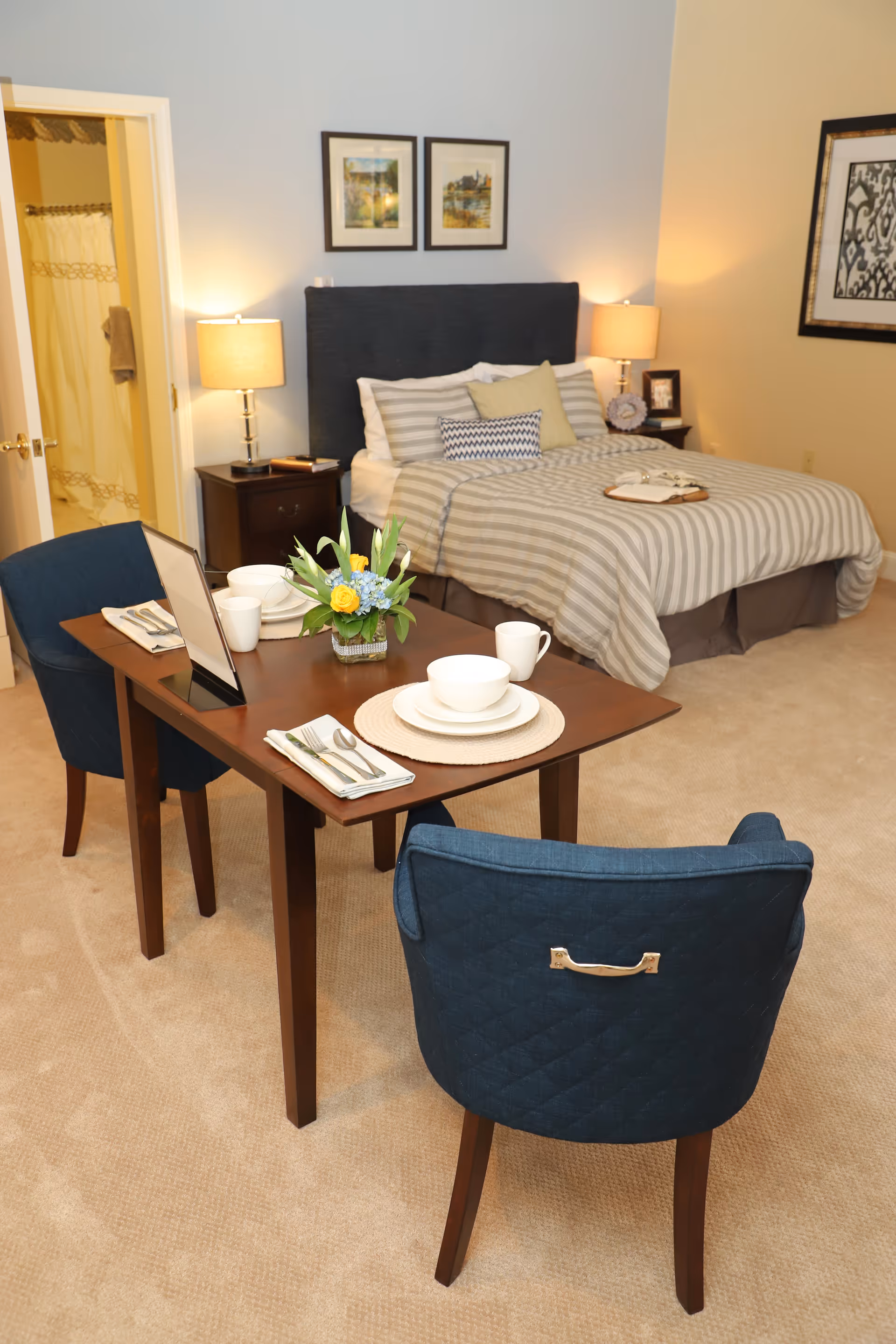 A furnished bedroom suite with a made bed, bedside lamps, and a small dining table set for two with blue chairs.
