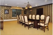 A formal dining room with a long dark wooden table surrounded by ten upholstered chairs with wooden frames. The room has patterned wallpaper, a beige carpet, a chandelier with multiple lights hanging above the table, and a framed picture on the wall.