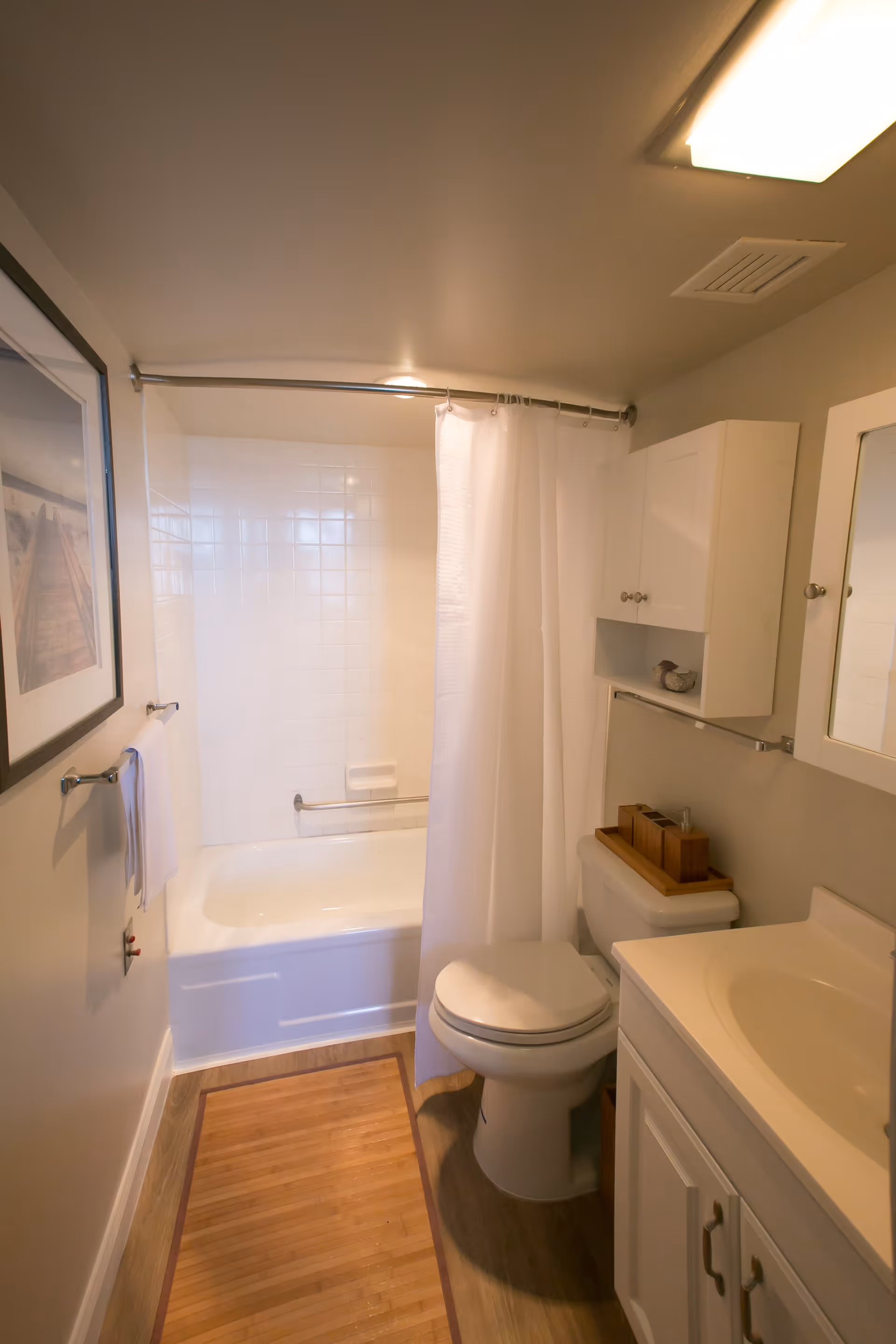 A small bathroom with a white bathtub and shower curtain, a toilet, a white vanity with a sink, a wall-mounted cabinet, a towel rack with a white towel, and a framed picture on the wall. The floor has a wooden mat and the walls are painted light beige.