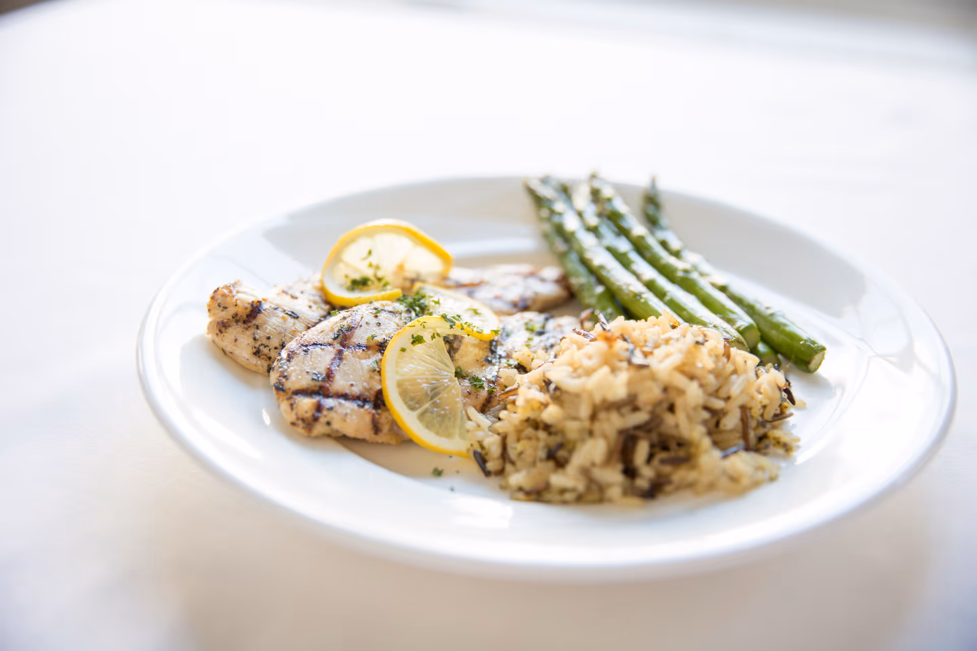 A white plate with grilled chicken breast garnished with lemon slices and herbs, served with a side of wild rice and steamed asparagus.