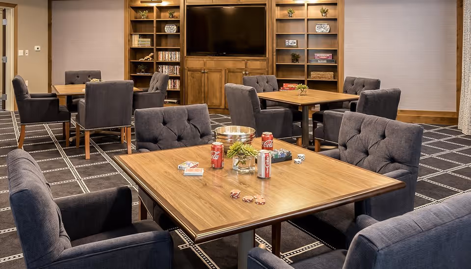 A cozy common area with multiple wooden tables surrounded by cushioned gray chairs. On the closest table, there are playing cards, poker chips, dice, three cans of Coca-Cola, a small plant, and a metal bowl. In the background, there are built-in wooden shelves with decorative items and a large flat-screen TV mounted above a wooden cabinet. The room has a patterned carpet and neutral-colored walls.