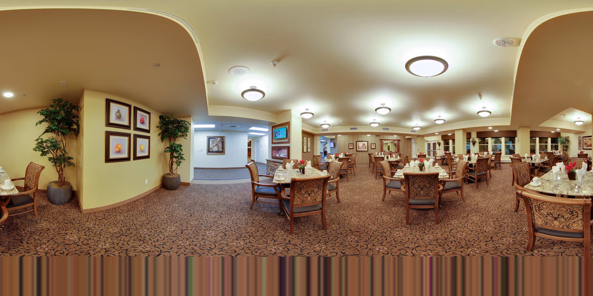 A spacious dining room in a senior living facility with multiple round tables set with white cups, saucers, and napkins. The room features patterned carpet, beige walls, framed artwork, and several potted plants. Ceiling lights provide warm illumination, and large windows line one side of the room.