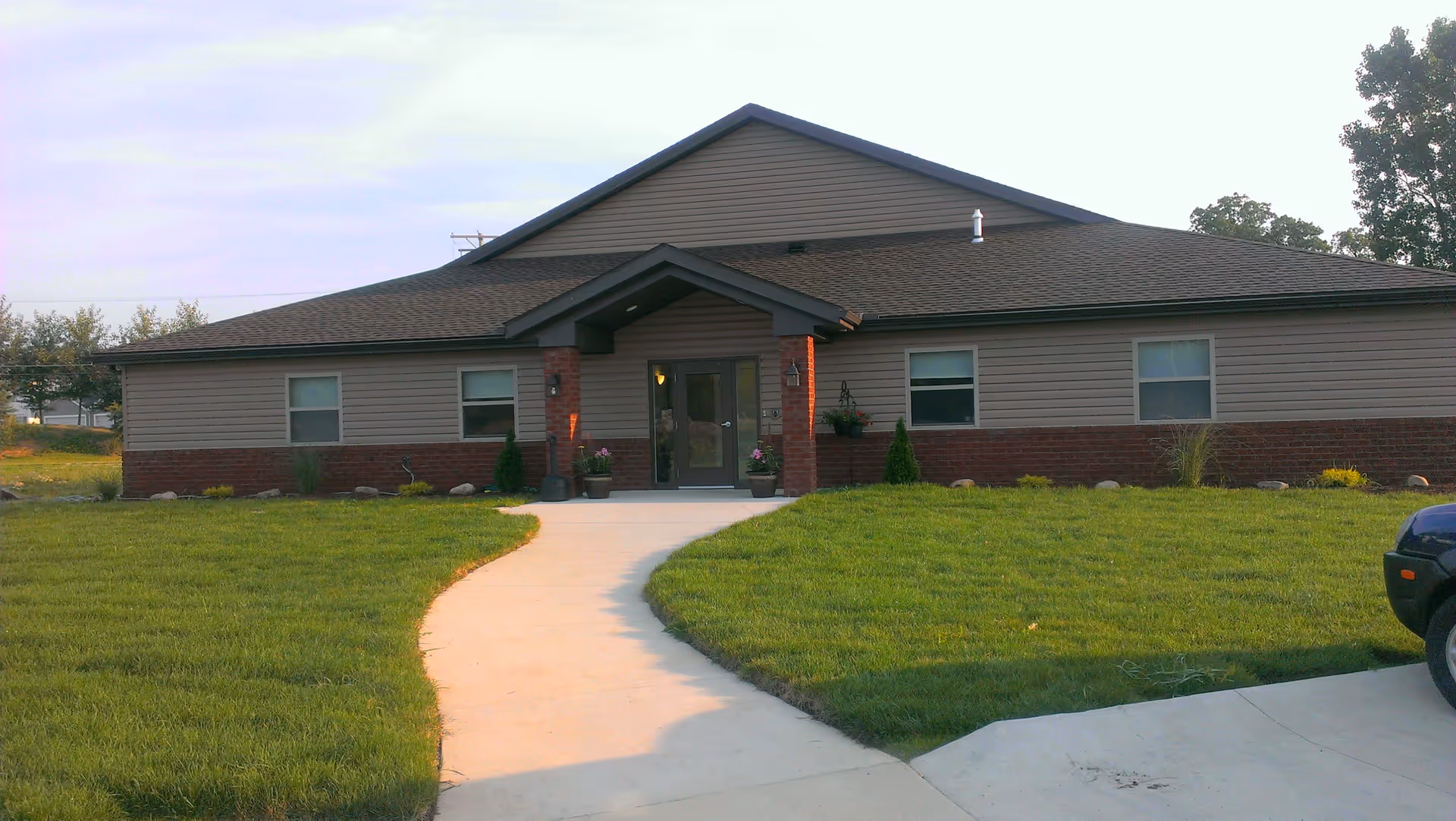 Front exterior view of a single-story senior care facility building with a sloped roof, brick and siding walls, a central entrance with a small covered porch, and a curved concrete walkway leading to the door. The building is surrounded by a well-maintained green lawn and some small shrubs and plants near the entrance.