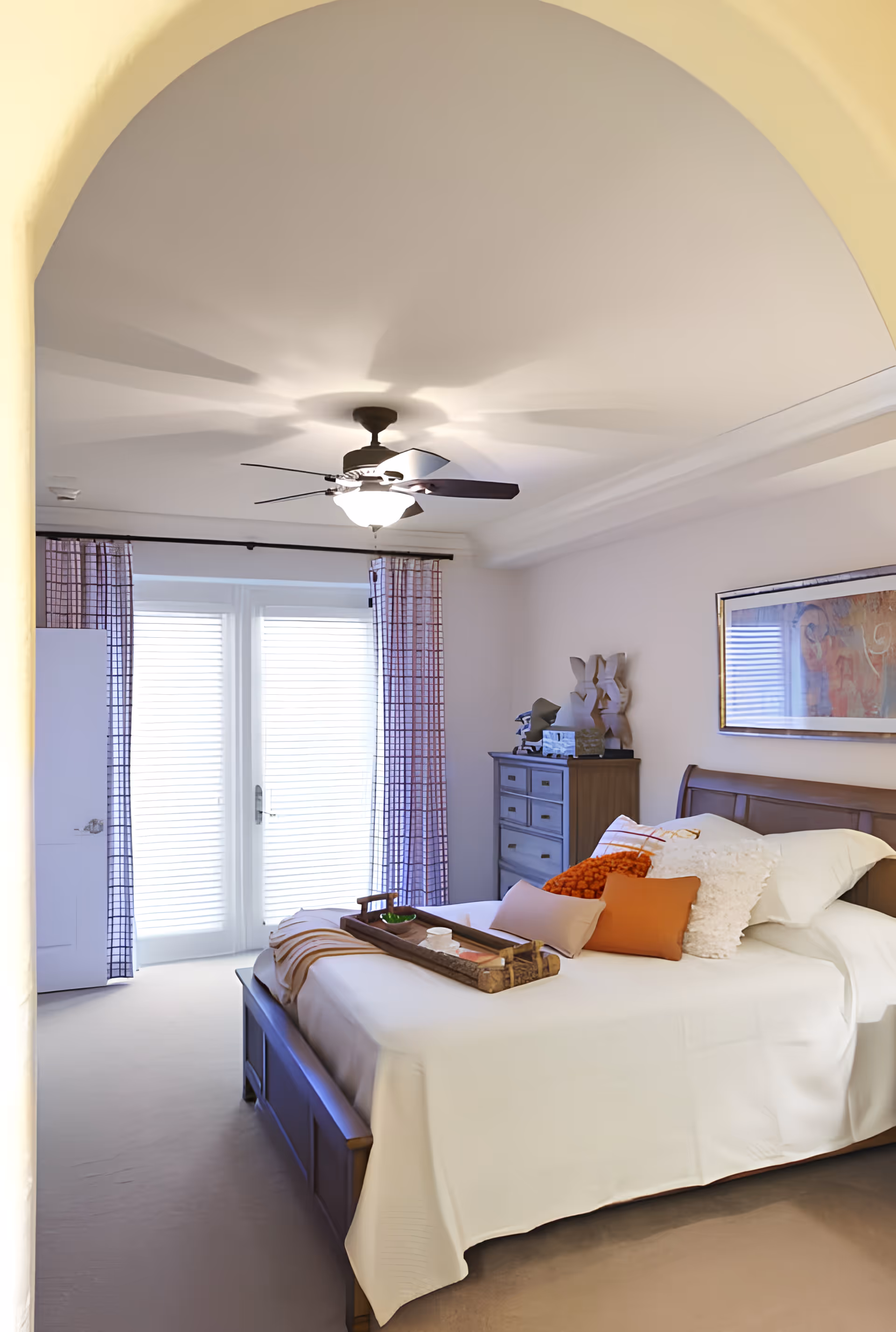 A bright, neatly made bedroom seen through an arched doorway, featuring a bed with decorative pillows, a ceiling fan, dresser, and glass doors with curtains.