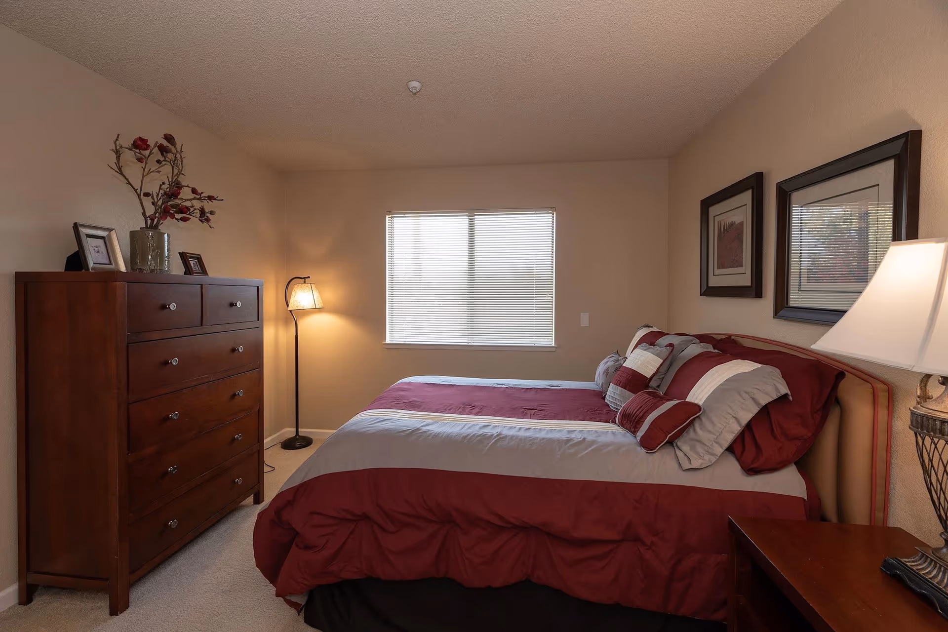A cozy bedroom with a large bed dressed in red and gray bedding, several pillows, a wooden dresser with decorative items on top, a floor lamp in the corner, and two framed pictures hanging on the wall above the bed. A window with blinds allows natural light into the room.