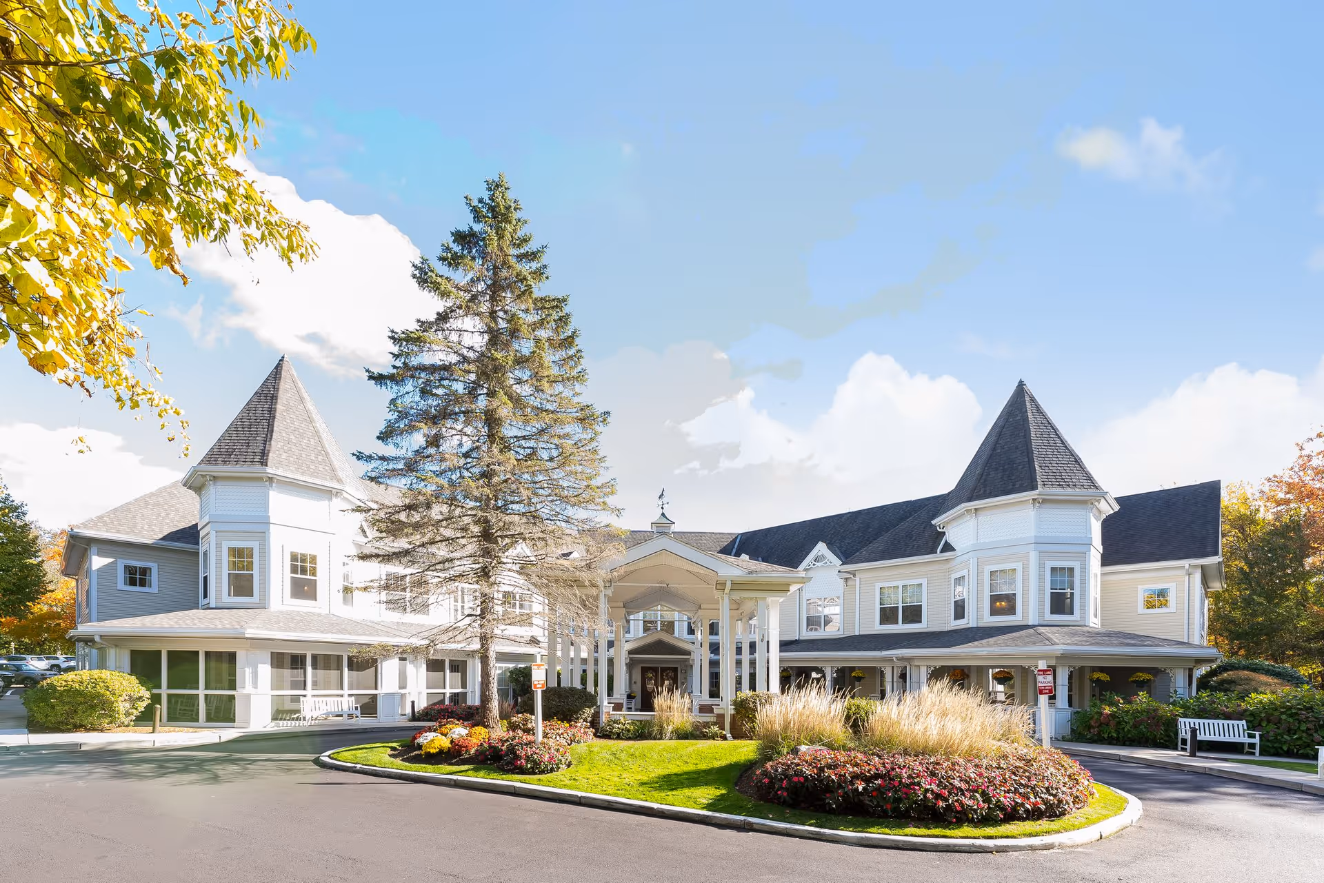 Exterior view of a large, elegant senior living facility building with two prominent turret-style roof sections, surrounded by landscaped greenery, flowers, and a paved driveway under a bright blue sky with some clouds.