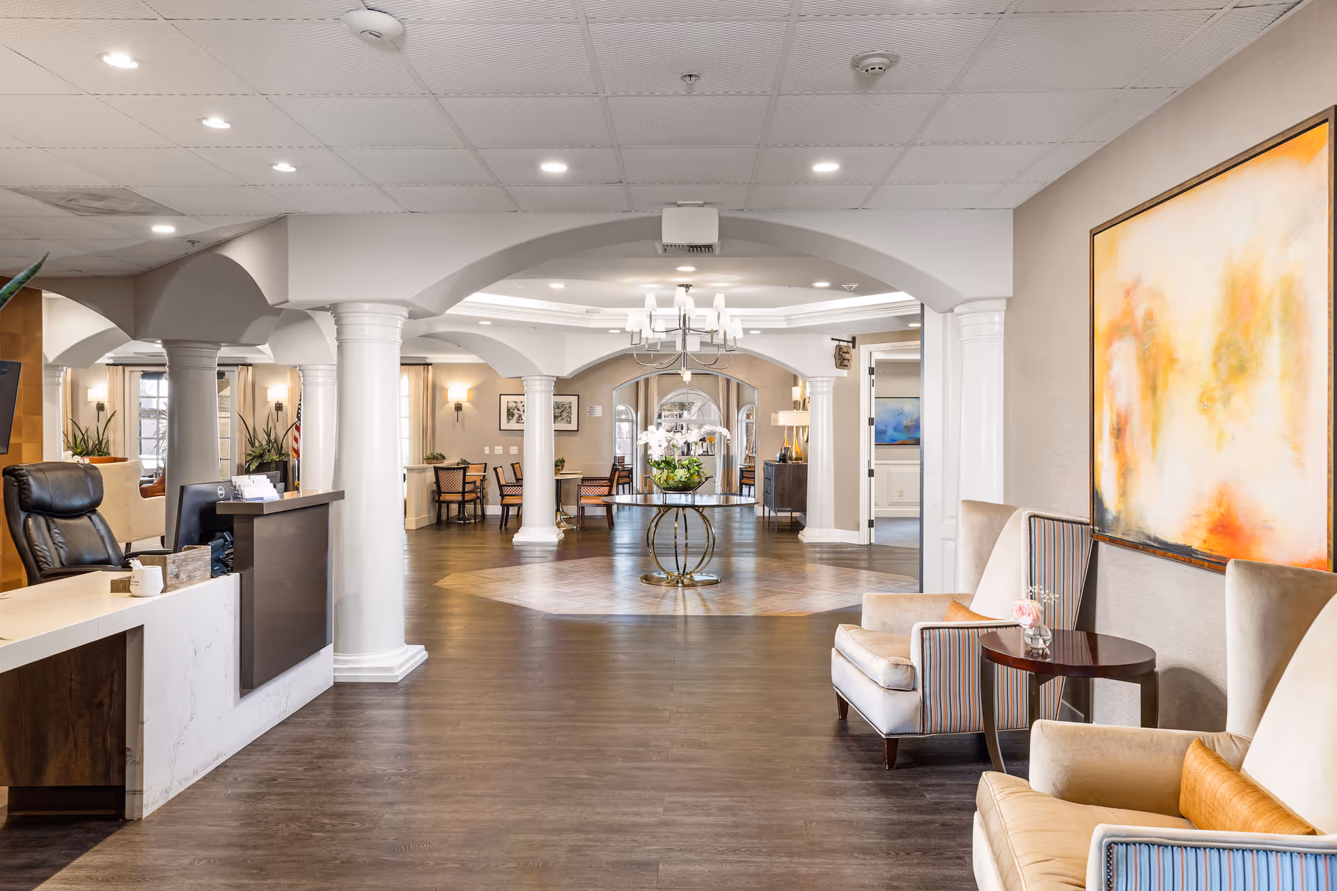 A spacious and elegant senior living facility lobby with white columns, a reception desk with a black leather chair, comfortable armchairs with striped upholstery and orange cushions, a round table with a flower arrangement in the center, and warm lighting. The walls are decorated with abstract paintings and framed pictures.