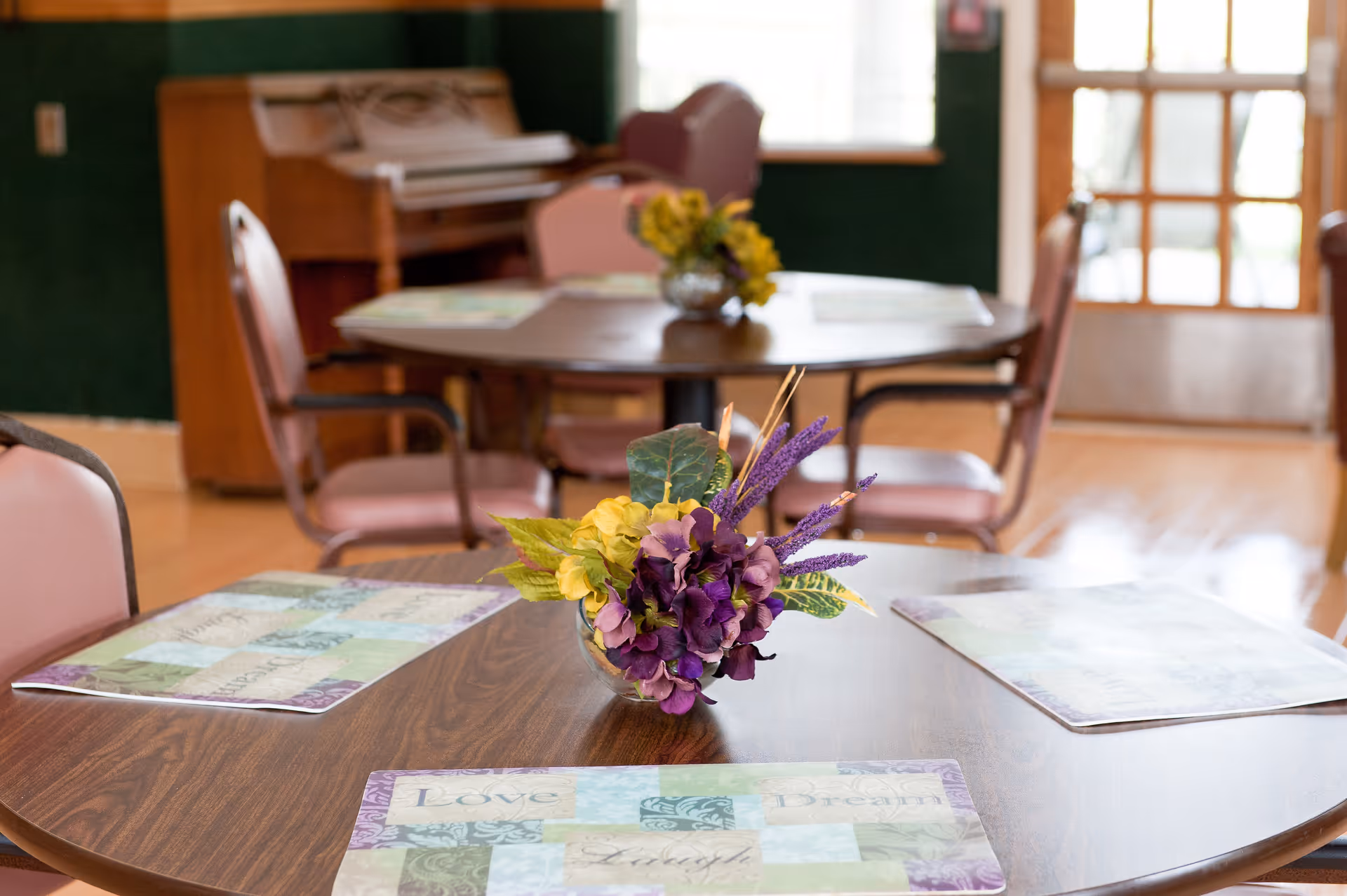 A cozy dining area with round wooden tables and pink cushioned chairs. Each table has a decorative floral centerpiece and placemats with words like 'Love' and 'Dream'. In the background, there is a piano and a door with glass panes letting in natural light.