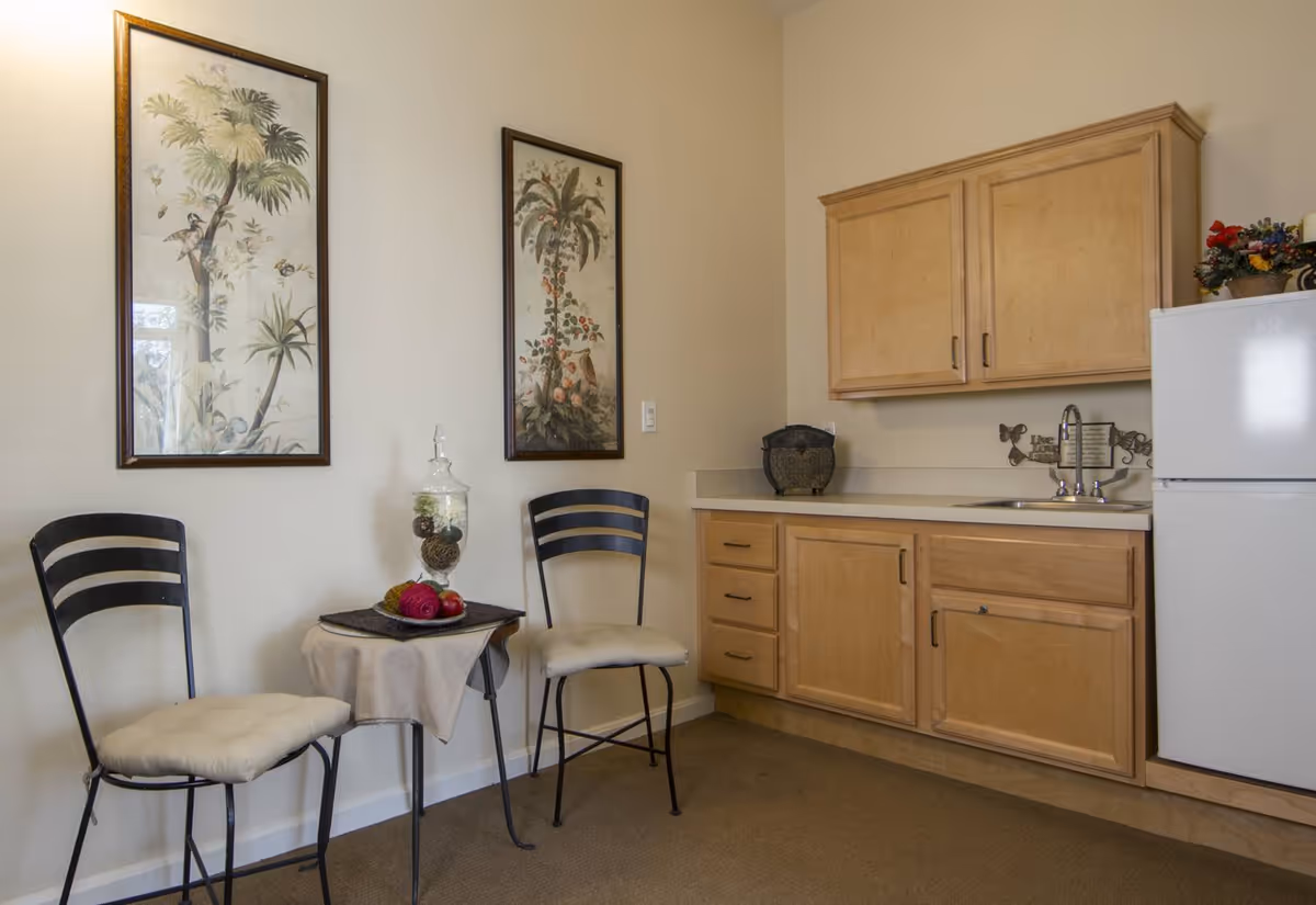 Small kitchenette area with light wood cabinets, a white refrigerator, and a sink. Two black metal chairs with white cushions are placed on either side of a small table covered with a beige cloth and decorative items. Two framed botanical prints hang on the wall above the chairs.