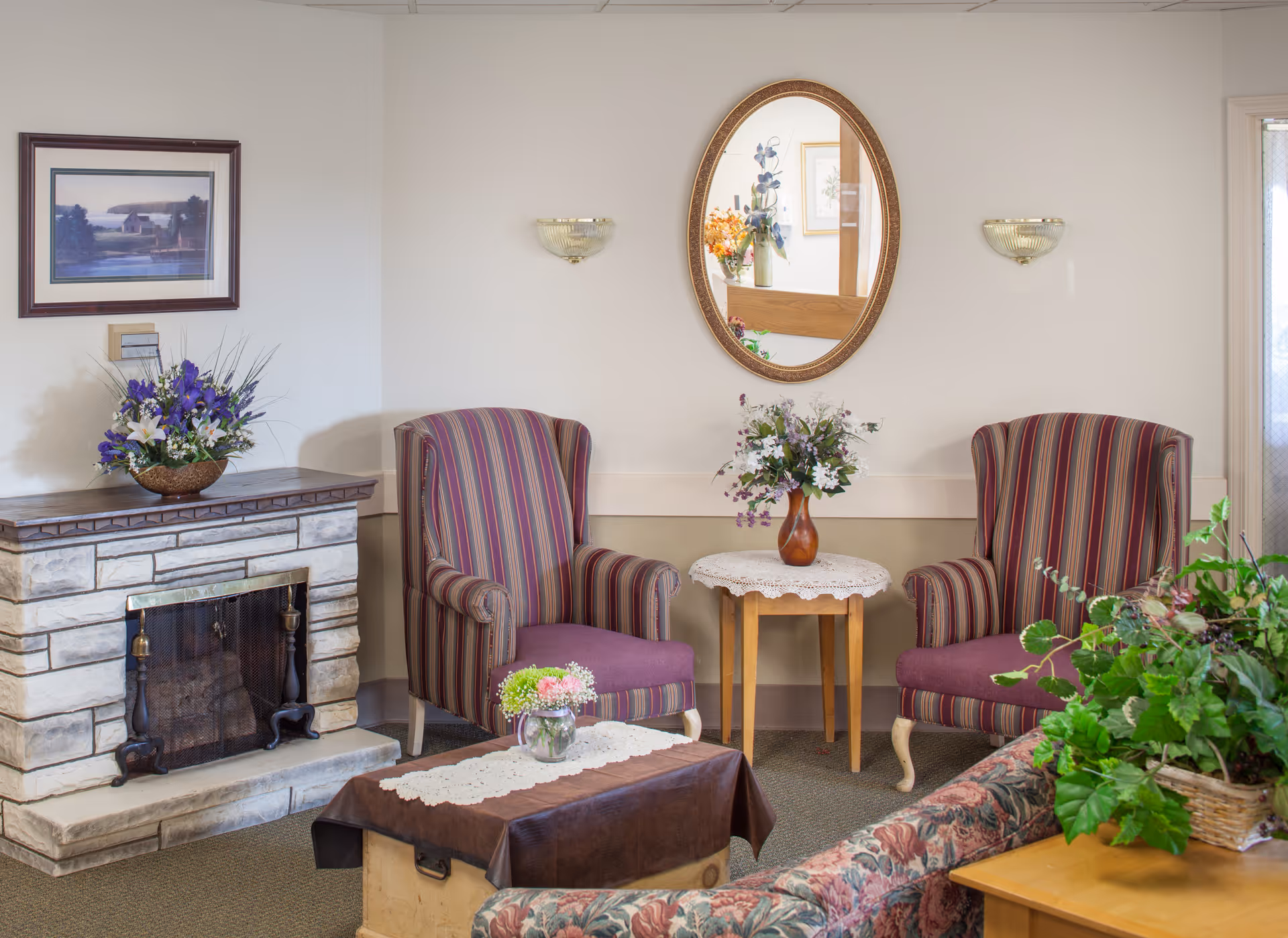 Cozy sitting area with two striped armchairs, a small table with flowers, a fireplace, and an oval mirror on the wall.