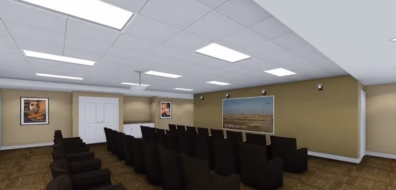A conference or presentation room with rows of black chairs facing a wall-mounted screen displaying a landscape image. The room has beige walls, a white ceiling with recessed lighting, and framed posters on the walls. There is a double door and a cabinet at the back of the room.