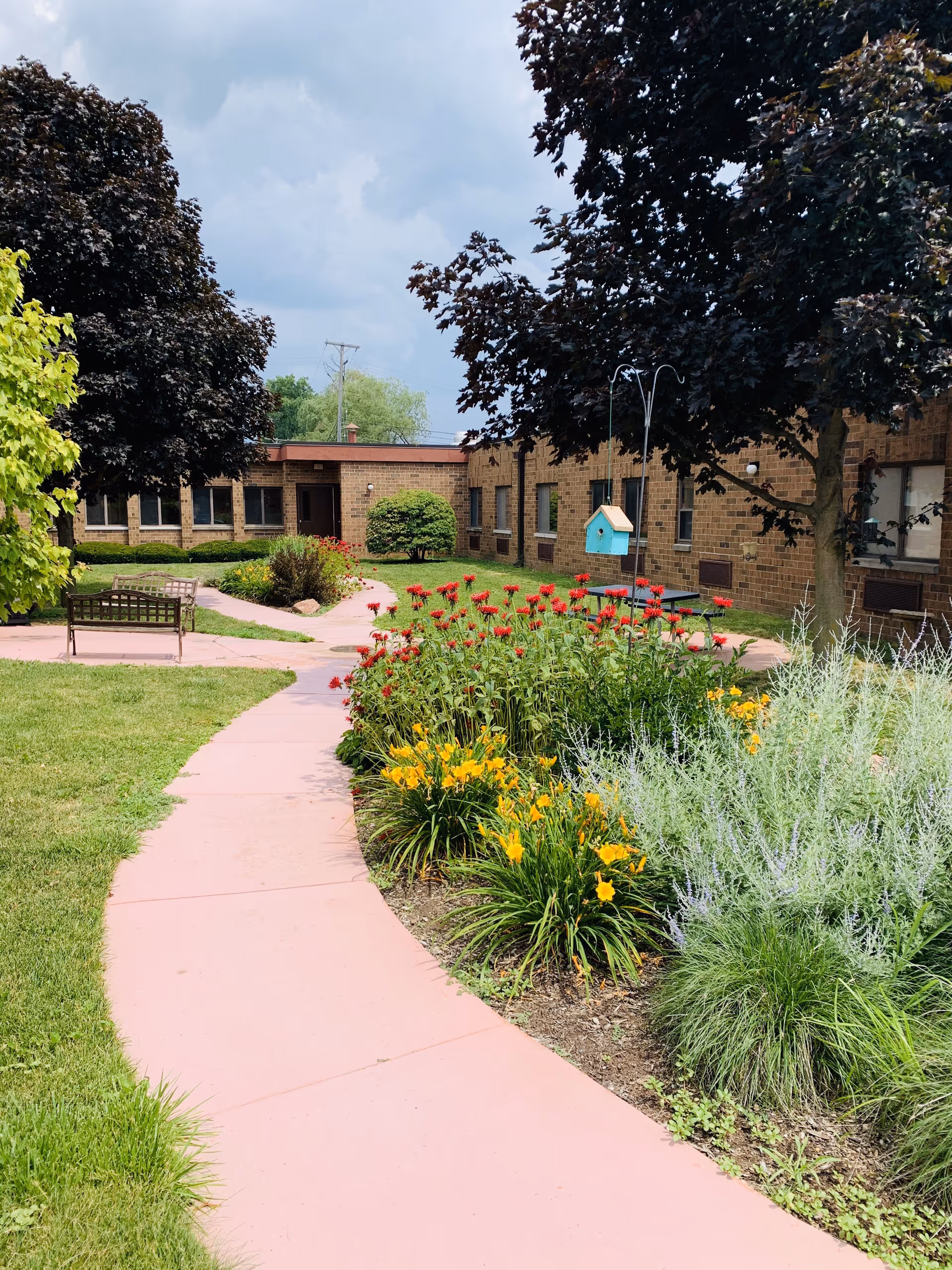 A landscaped outdoor garden area with a pink paved walkway curving through green grass and colorful flower beds with red and yellow flowers. There are benches along the path and a blue birdhouse hanging from a metal pole. The background shows a single-story brick building under a partly cloudy sky.
