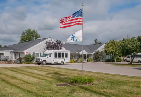 Exterior view of Carriage Court of Marysville facility with a white building, a small shuttle bus parked in front, and two flagpoles displaying the American flag and a white flag with a blue logo. The scene includes a well-maintained lawn, trees, and a partly cloudy sky.