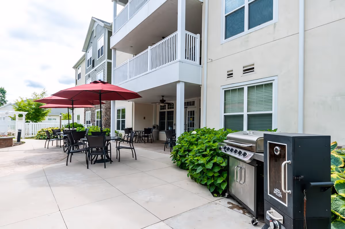 Outdoor patio area at Arbor Terrace Willistown with tables and chairs under red umbrellas, a barbecue grill, and a smoker next to a building with balconies and windows.