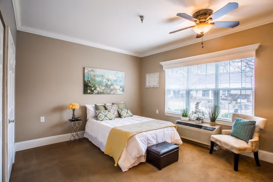 A cozy bedroom with a neatly made bed featuring white and yellow bedding and decorative pillows. There is a small side table with a vase of yellow flowers next to the bed. A cushioned chair with a green pillow is placed near a large window with blinds, allowing natural light to fill the room. The walls are painted beige, and a ceiling fan with a light fixture is mounted on the ceiling. A piece of floral artwork hangs above the bed.