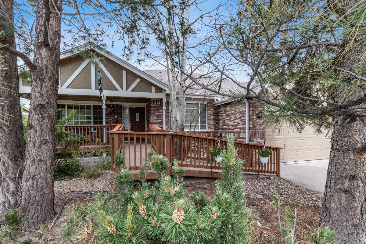 Front exterior view of a single-story brick house with a wooden ramp leading to the front door, surrounded by trees and shrubs under a clear blue sky.
