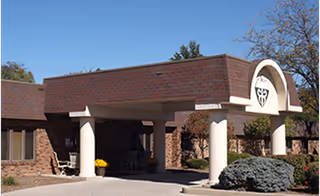 Exterior view of a brick building with a covered entrance supported by white columns. There is a decorative emblem above the entrance and some landscaping including bushes and a tree with autumn leaves.
