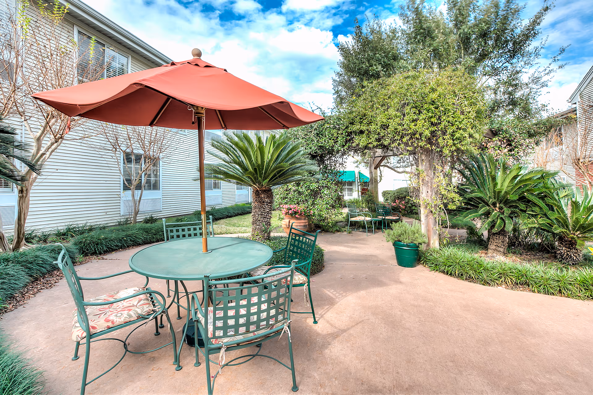 Outdoor patio area with a round green metal table and four matching chairs, one with a floral cushion, under a large red umbrella. The patio is surrounded by greenery including palm plants, bushes, and a wooden pergola covered with vines. The exterior of a light-colored building with windows is visible in the background under a partly cloudy sky.