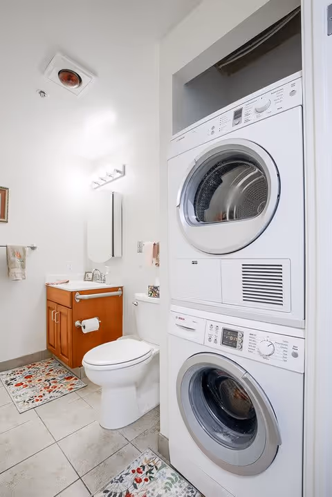 A bathroom with a white toilet, wooden vanity with a sink, mirror, and light fixture above. There is a stacked washer and dryer unit on the right side. The floor has light-colored tiles with floral-patterned rugs.