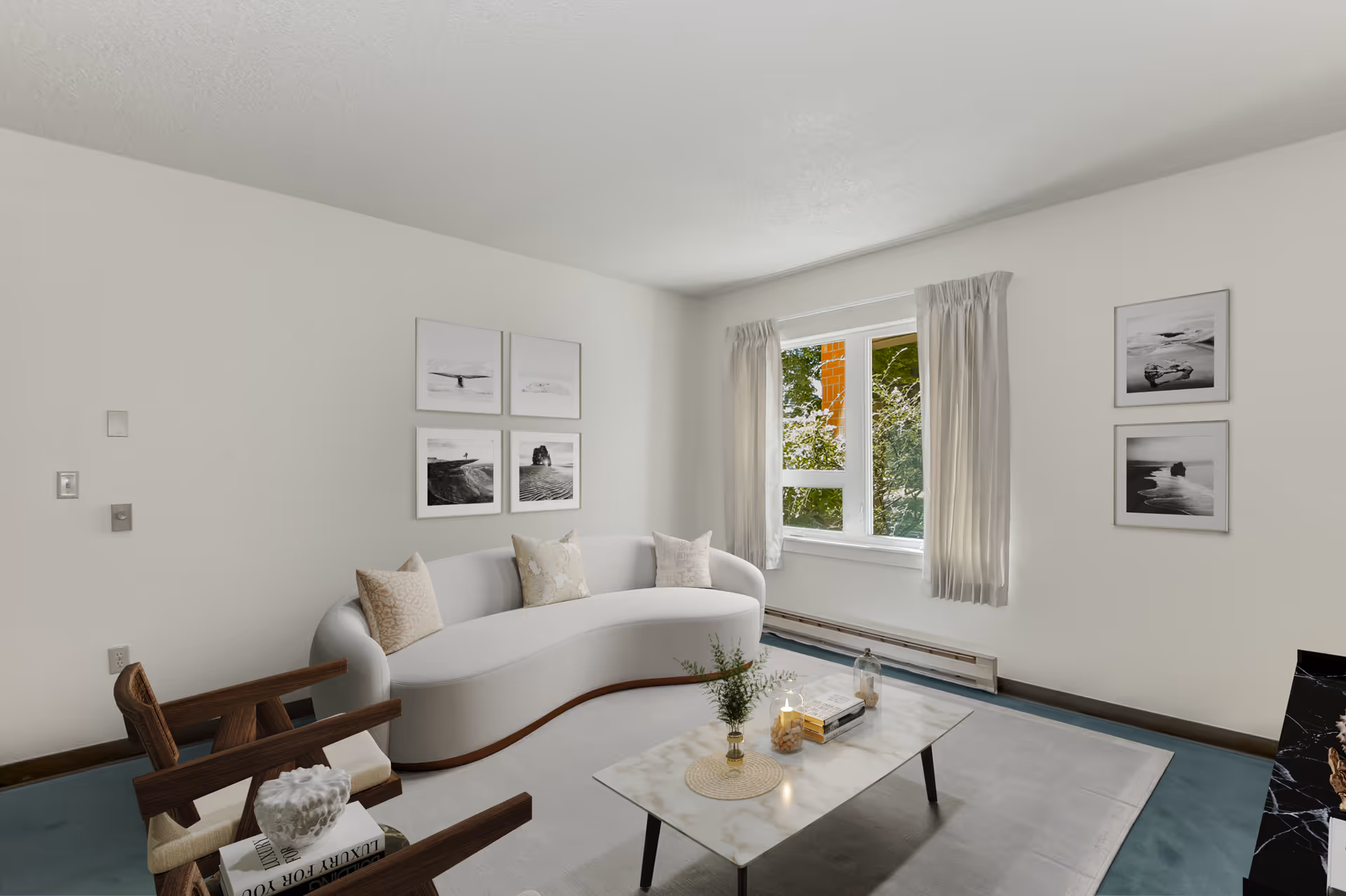 A bright living room with a curved white sofa adorned with three pillows, a wooden armchair with a cushion, and a rectangular marble coffee table holding decorative items. The room has light-colored walls with six black and white framed photographs, a window with light curtains, and a blue carpeted floor.