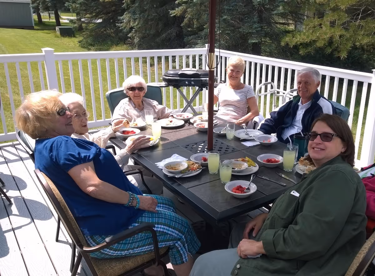 Six older adults sitting around a patio table on a sunny deck enjoying food and drinks.