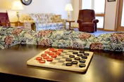 A cozy living room area in Oakview Nursing Home featuring a checkers board with red and black pieces on a dark wooden table. In the background, there is a floral-patterned sofa, a brown armchair, a side table with a lamp, and a wall clock.