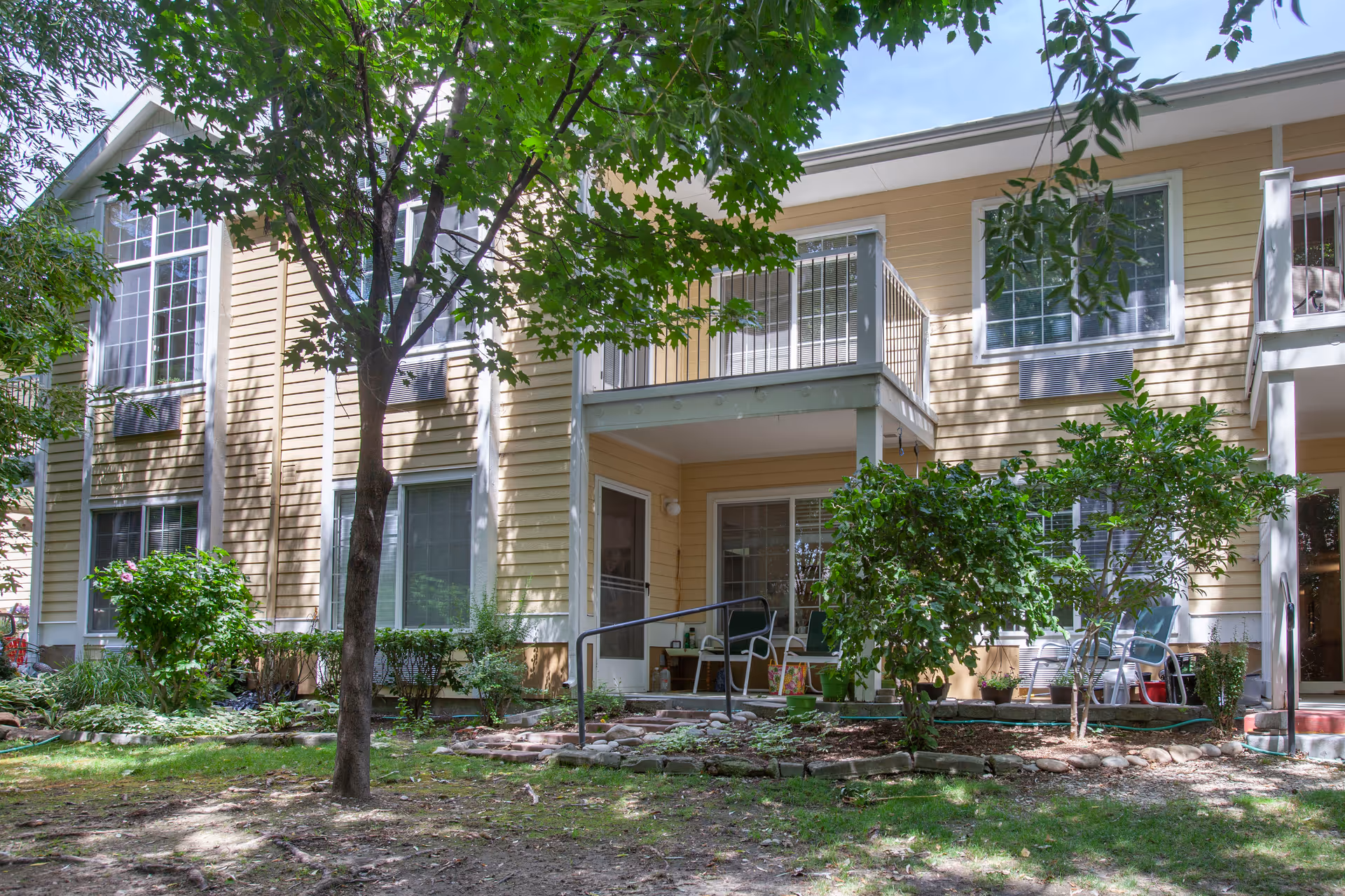 Two-story yellow apartment building with balconies and ground-level patios surrounded by trees and landscaping.