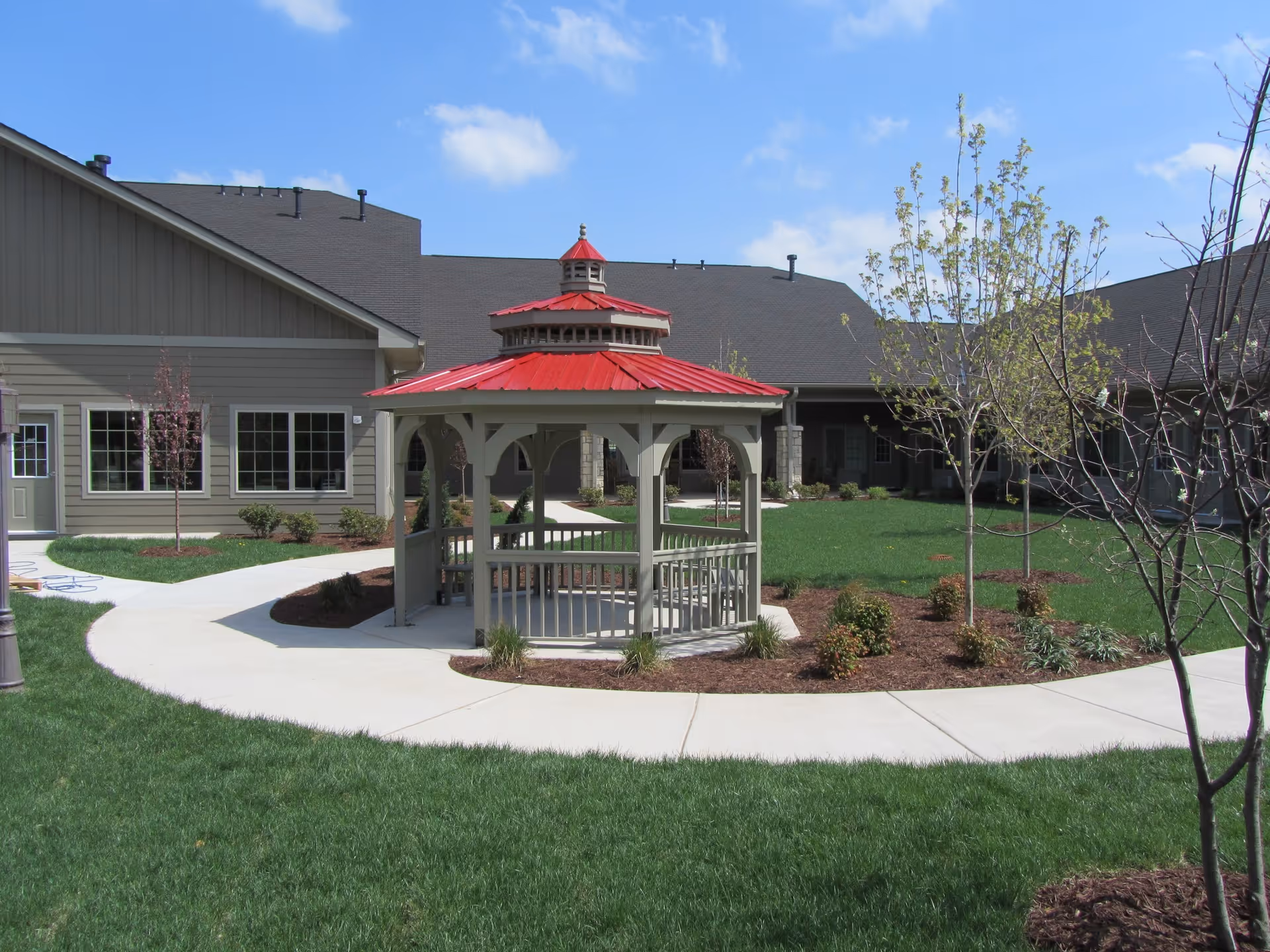 Outdoor courtyard area of a senior living facility with a central gazebo featuring a red roof, surrounded by a curved concrete walkway, green grass, young trees, and shrubs. The building with gray siding and multiple windows is visible in the background under a blue sky with some clouds.