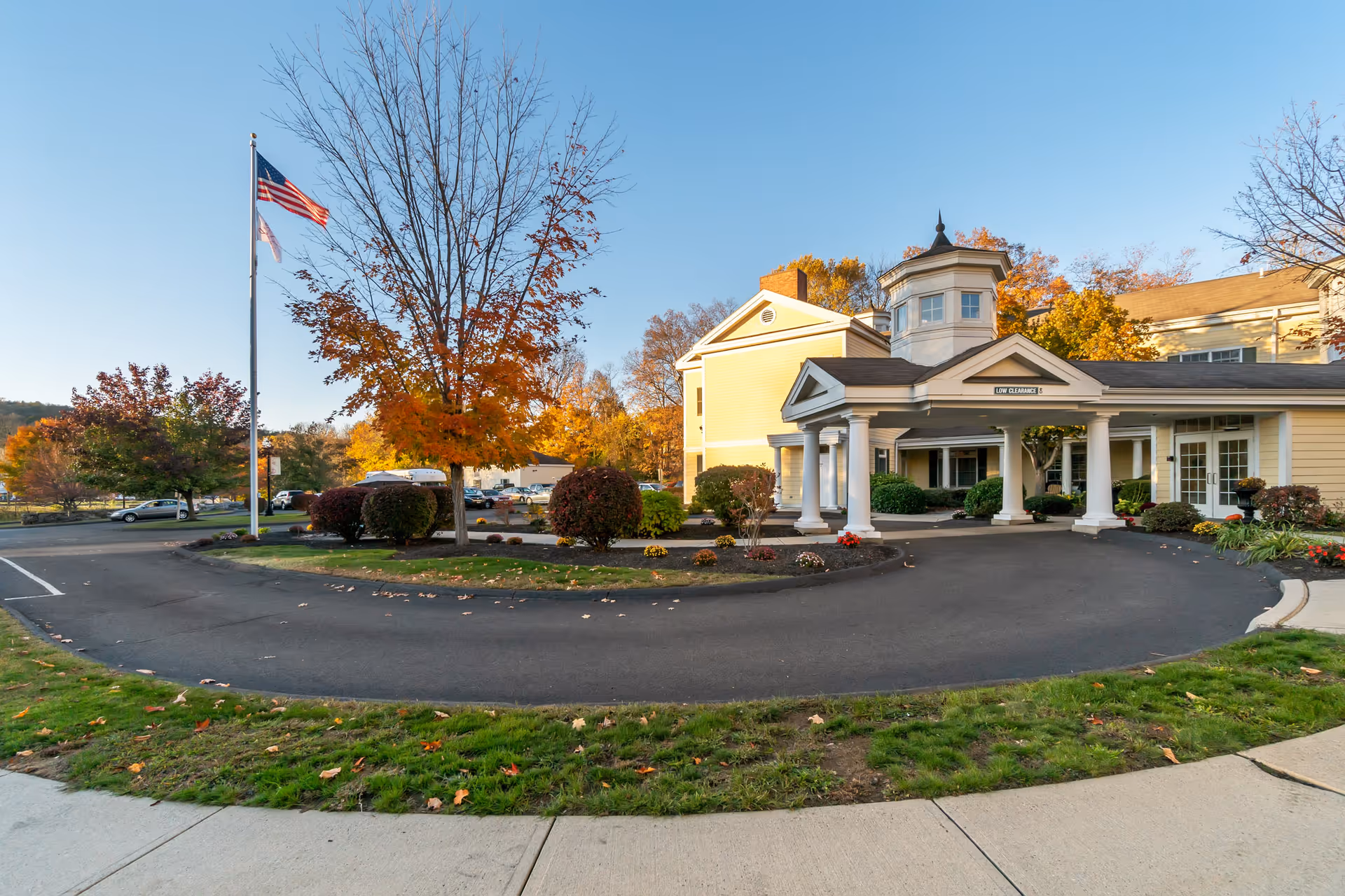 Exterior view of Monarch Southbury facility with a circular driveway, landscaped bushes, and trees with autumn foliage. An American flag is flying on a flagpole near the driveway entrance. The building is light yellow with white columns and a covered entrance.