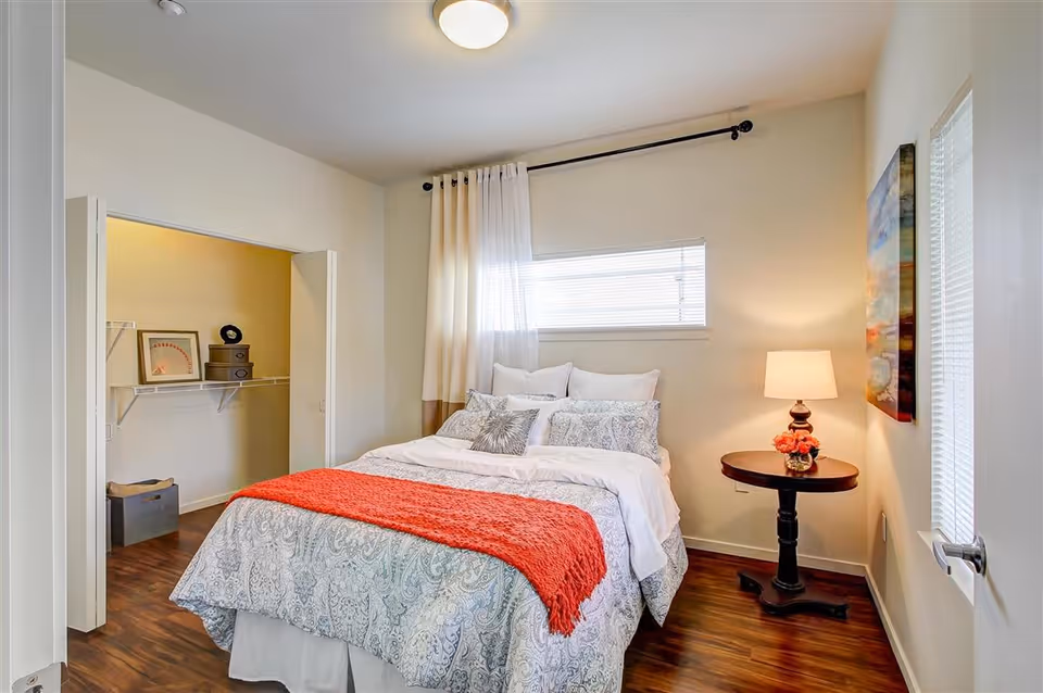A cozy bedroom with a neatly made bed featuring patterned bedding and a red throw blanket. There is a small round wooden side table with a lamp and a vase of flowers. The room has wooden flooring, a window with blinds, and an open closet with shelves and storage boxes.