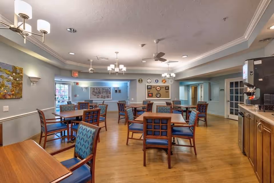 A well-lit dining room with multiple wooden tables and chairs featuring blue patterned cushions. The room has light blue walls, wooden flooring, ceiling fans, and several ceiling lights. There is a coffee machine on a counter to the right and artwork on the walls.