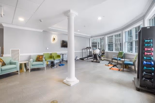 A bright and spacious senior living fitness room with exercise equipment including a treadmill, stationary bike, dumbbells, and resistance bands. The room has large windows letting in natural light, light gray walls, and comfortable seating with green and blue chairs along the walls. A white column stands in the center of the room.