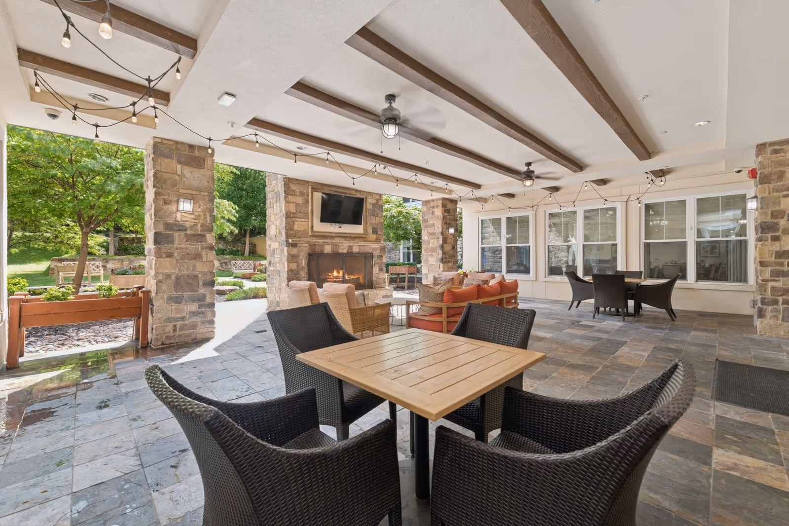 Covered outdoor seating area with stone pillars and ceiling beams, featuring a fireplace with a mounted TV above it. There are several tables with wicker chairs and a seating area with orange cushions. The space opens to a garden with trees and benches.