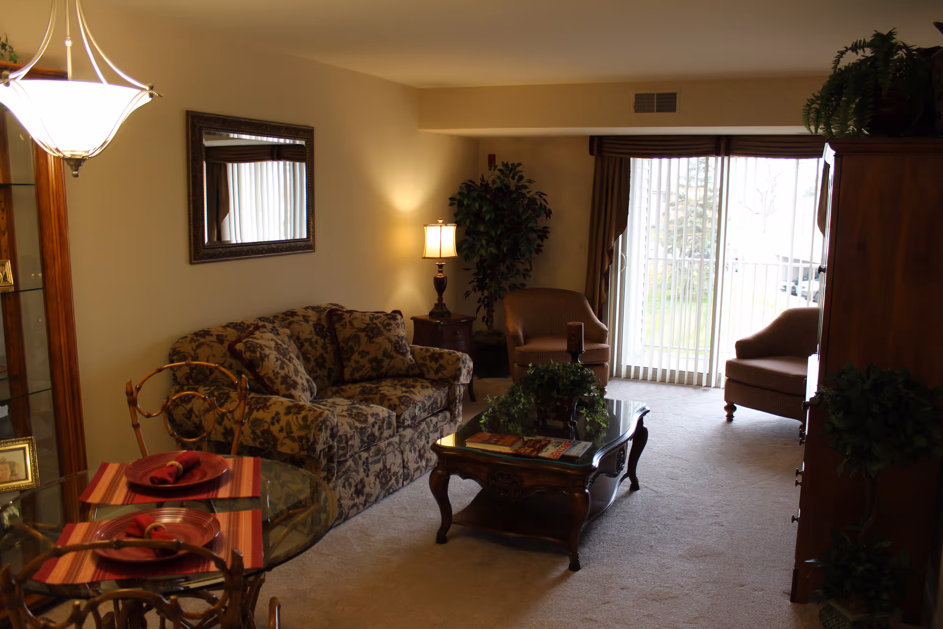 A cozy living room area with a floral patterned sofa, two armchairs, a wooden coffee table with plants and magazines, a side table with a lamp, a large mirror on the wall, and sliding glass doors with vertical blinds leading outside. A small glass dining table with two place settings is visible in the foreground.