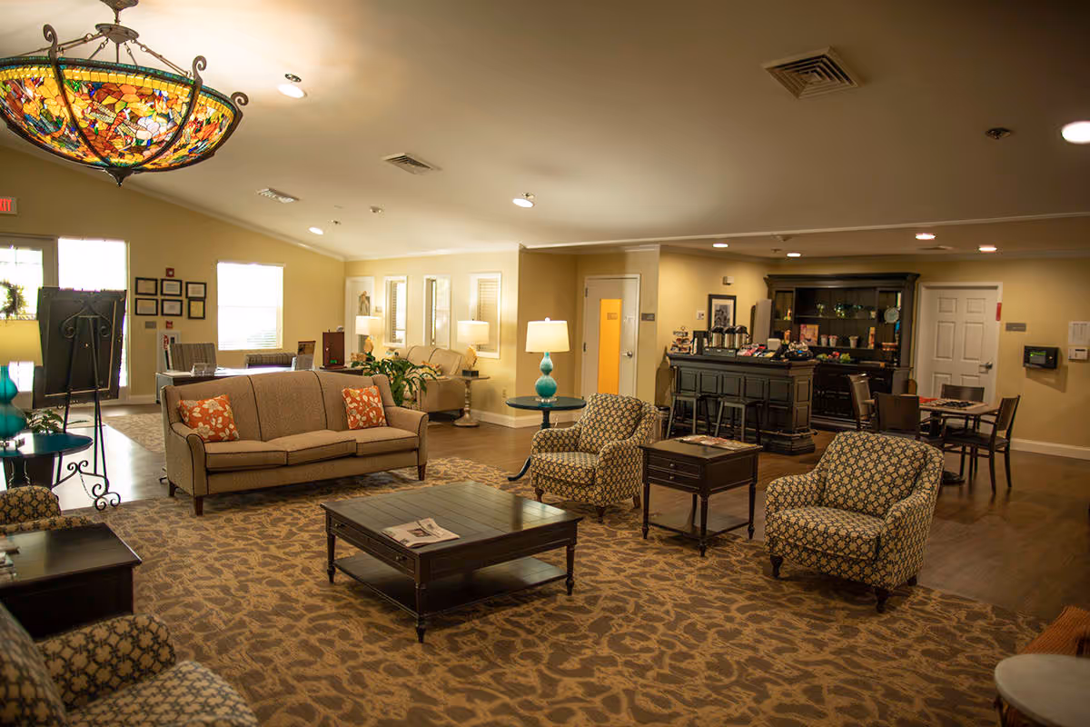 A spacious and well-lit living room area in a senior living facility featuring a beige sofa with patterned cushions, two patterned armchairs, a dark wooden coffee table, side tables with lamps, and a carpet with a floral design. In the background, there is a counter area with coffee and refreshments, additional seating with tables and chairs, and large windows letting in natural light.