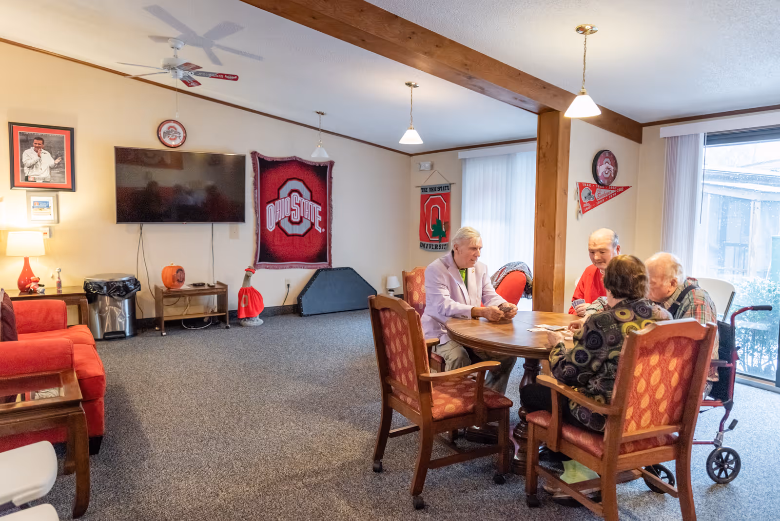 A group of four elderly people sitting around a wooden round table playing cards in a cozy living room area. The room has carpeted floors, a red couch, a wall-mounted TV, and Ohio State University decorations including a wall hanging, pennants, and framed pictures. Large windows with vertical blinds allow natural light into the room.