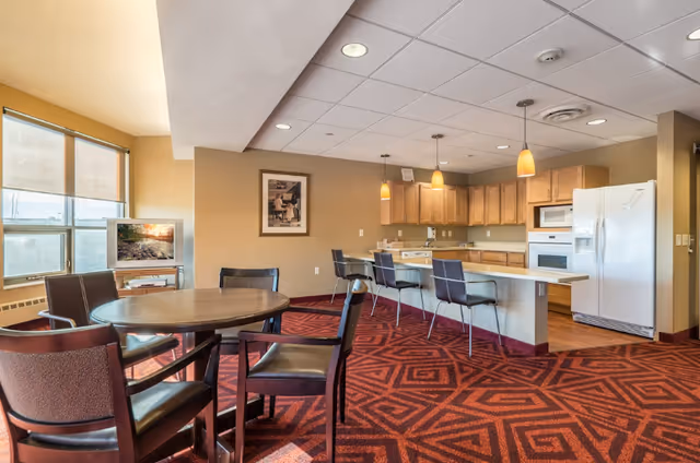 Communal dining area and kitchenette with a round dining table, chairs, bar stools at a counter, and large windows.