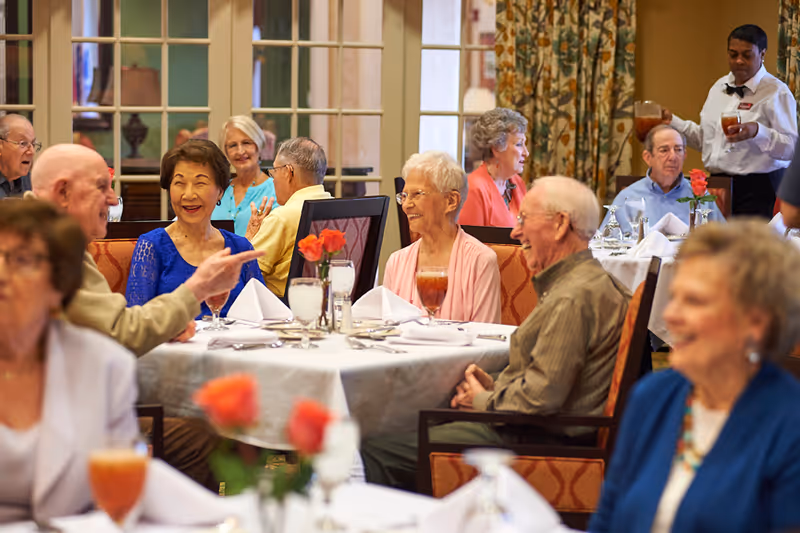 A group of elderly people sitting at tables in a dining room, enjoying conversation and drinks. A server is pouring a beverage at one of the tables. The tables are set with white tablecloths, napkins, glasses, and small vases with red flowers. The room has large windows with patterned curtains.