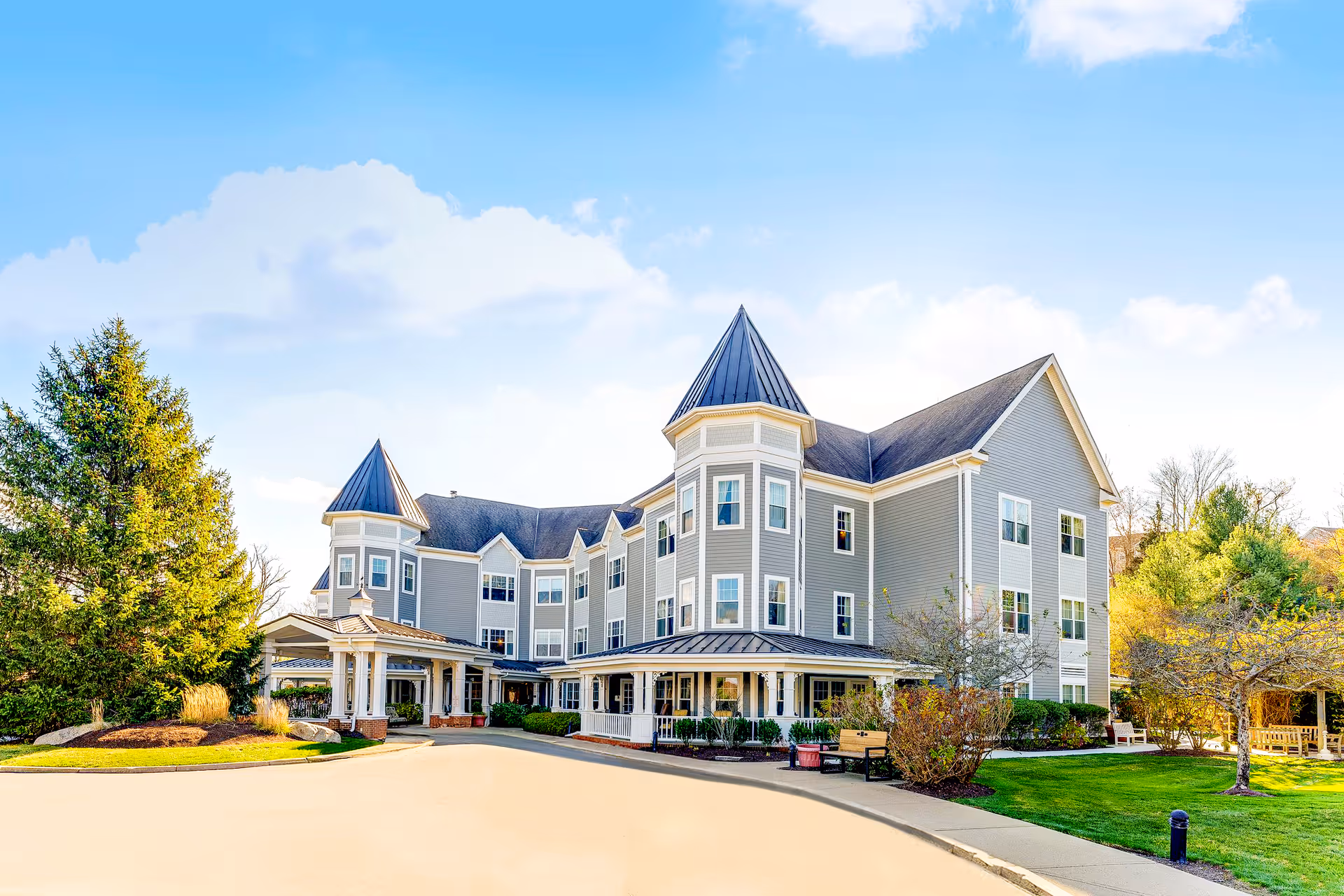 Exterior view of a large, three-story senior living facility building with gray siding and multiple peaked roofs. The building is surrounded by landscaped greenery, including trees and bushes, with a clear blue sky overhead. There is a driveway and a covered entrance area in front of the building.
